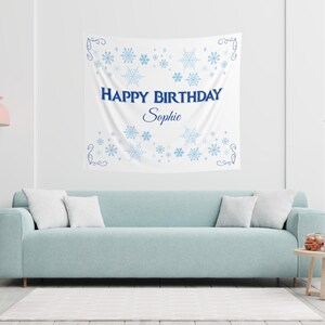 Puede incluir: Un tapiz de pared blanco con copos de nieve azules y el texto "HAPPY BIRTHDAY Sophie". Se muestra sobre un sof&aacute; azul claro con cojines blancos. Tambi&eacute;n se ven una l&aacute;mpara rosa y una mesa auxiliar.