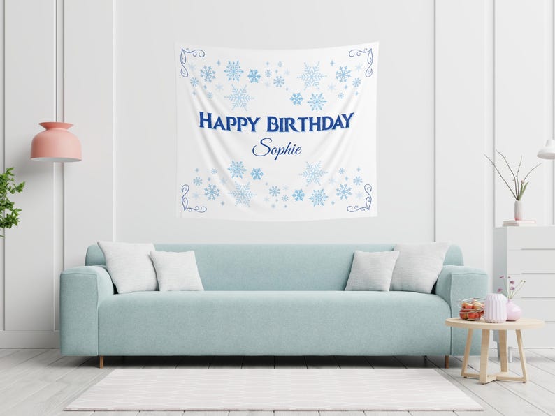 Puede incluir: Un tapiz de pared blanco con copos de nieve azules y el texto "HAPPY BIRTHDAY Sophie". Se muestra sobre un sof&aacute; azul claro con cojines blancos. Tambi&eacute;n se ven una l&aacute;mpara rosa y una mesa auxiliar.