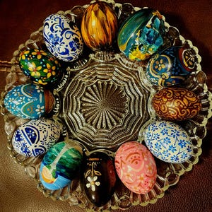 May include: A selection of twelve hand-painted Easter eggs, each with unique designs and colours, displayed on a clear glass plate. The eggs showcase intricate patterns, such as floral and abstract designs. The colour scheme includes blue, green, yellow, brown, and pink.