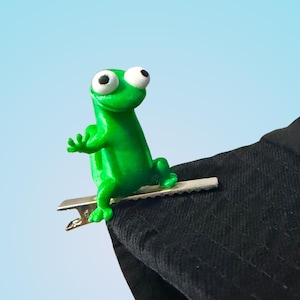 May include: A vibrant green frog ornament with big white eyes and a silver clip, attached to the edge of a black hat. The frog is positioned on the hat, with its arms extended. The background is a light blue gradient.