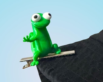 May include: A bright green frog figurine with large white eyes and a silver clip, attached to the edge of a black hat. The frog is perched on the hat, with its arms outstretched. The background is a gradient of light blue.