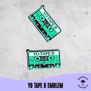 Puede incluir: Dos emblemas de cinta de cassette verde y negro con el texto "YO TAPE B". Los emblemas están sobre un fondo gris claro. La parte inferior de la imagen tiene el texto "YO TAPE B EMBLEM".