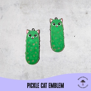 May include: Two green pickle-shaped cat emblems with pink ears and smiling faces. The pickles have brown spots and a small hole at the top. The text "PICKLE CAT EMBLEM" is at the bottom of the image.