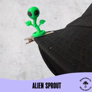 Könnte beinhalten: Ein grüner Alien-Spross mit schwarzen Augen und einem pflanzenartigen Körper ist an einem schwarzen Hut befestigt. Der Alien ist an einer silbernen Klammer befestigt. Der Text "ALIEN SPROUT" wird auf einem lila Banner angezeigt.