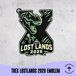 May include: A black and green emblem featuring a T-Rex head with an open mouth, positioned over a large letter X. The text LOST LANDS 2026 is printed in white at the bottom. The design includes a small circular loop at the top for hanging.