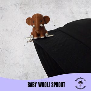 Puede incluir: Una pinza para el pelo con forma de elefante marrón con detalles blancos, sujeta a una tela negra. La pinza es plateada. El texto "BABY WOOLI SPROUT" está en la parte inferior de la imagen.