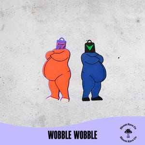 Puede incluir: Dos figuras de dibujos animados, una naranja y otra azul, con los brazos cruzados y una cabeza en forma de bolsa. La figura naranja tiene una cabeza morada, y la figura azul tiene una cabeza negra con una marca de verificación verde. El texto "WOBBLE WOBBLE" está en la parte inferior.