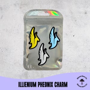 以下が含まれることがあります： 3つのIllenium Pheonixチャームが入った透明な虹色のパッケージ。チャームは、黄、水色、白のスタイリッシュな鳥のデザインで、黒いアウトラインが特徴です。「ILLENIUM PHEONIX CHARM」の文字が下部に印刷されています。