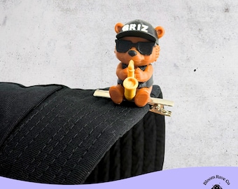May include: A novelty hair clip featuring a brown bear wearing sunglasses and a black cap with the word "GRIZ". The bear is holding a yellow saxophone. The clip is attached to a black, textured fabric.
