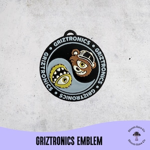 May include: A black and white emblem with the text "Griztronics" encircling a yin-yang design. One side features a cartoon bear wearing a hat, and the other side has a yellow monster with an eye. The text "Griztronics Emblem" is at the bottom.