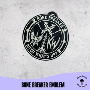 May include: A circular black and white emblem featuring a skull with wings, a spinal column, a cross, and the letters KW. The text BONE BREAKER and BITCH WHAT'S UP? is printed along the border. The design is framed by a bone-shaped circular border.