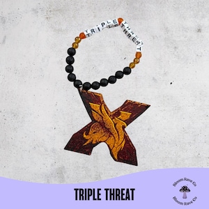 May include: A bracelet with the words "TRIPLE THREAT" spelled out in white letter beads. The bracelet features black lava beads, orange and yellow beads, and a wooden pendant in the shape of an "X" with a gold design.