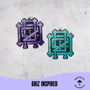 May include: Two decorative pendants with a black background and a stylized design. One is purple, the other is teal. The pendants have a small silver loop at the top for hanging. The text "GRIZ INSPIRED" is at the bottom.
