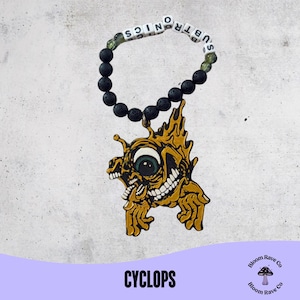 Peut inclure: Un bracelet de perles noires avec le mot "SUBTRONICS" épelé en perles de lettres blanches. Un pendentif cyclope de couleur or avec un grand œil et un sourire édenté est suspendu au bracelet. Le mot "CYCLOPS" est imprimé en blanc sur une bannière violette.