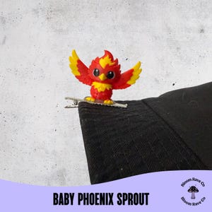 May include: A red and yellow baby phoenix sprout hair clip. The clip is attached to a black fabric. The phoenix has outstretched wings and a detailed design. The text "BABY PHOENIX SPROUT" is at the bottom of the image.