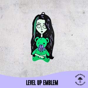 Puede incluir: Un emblema negro y verde con una ilustración estilizada de una niña con pelo largo negro y cara verde, sosteniendo un oso de peluche verde con un lazo morado. El texto "LEVEL UP EMBLEM" está en la parte inferior.