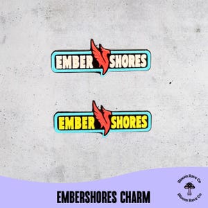 May include: Two rectangular enamel pins featuring the text Ember Shores in a bold font. The top pin has cream-colored lettering on a light blue background, while the bottom pin has yellow lettering. Both feature a red phoenix icon.