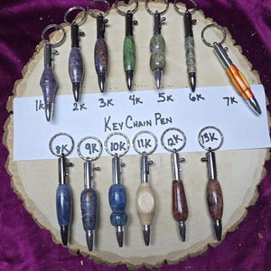 May include: A collection of keychain pens with various wood grain patterns and colors, including purple, green, and brown. Each pen has a silver-colored metal tip and keyring. The text "Key Chain Pen" is visible.