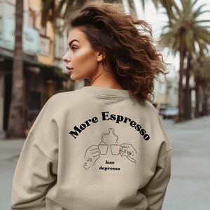 Op de afbeelding: Beige sweatshirt met de zwarte tekst "More Espresso less depresso". Het ontwerp bevat een lijntekening van twee handen die koffiekopjes vasthouden. De sweatshirt heeft een neutrale kleur en een relaxte pasvorm.