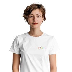 Puede incluir: Camiseta blanca con la palabra "love" en cursiva y corazones de colores del arcoíris. La camiseta es de estilo cuello redondo clásico y está hecha de un material suave y cómodo. El diseño está situado en la parte superior izquierda.
