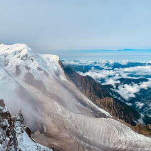 May include: A panoramic vista of snow-capped mountains and a glacier, with clouds filling the valley. The sky is a clear blue, creating a sense of scale and natural beauty.