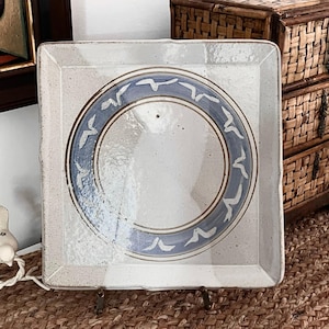 May include: Square ceramic plate with a speckled, off-white surface. A blue ring with white bird-like shapes encircles the center. The plate has a slightly raised edge and a rustic, handmade appearance.