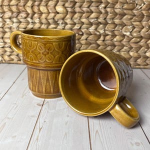May include: Two stacked, vintage-style, brown ceramic mugs with handles. The top mug features a textured pattern of circles and arches, while the bottom has a geometric design. The mugs are on a light wood surface.