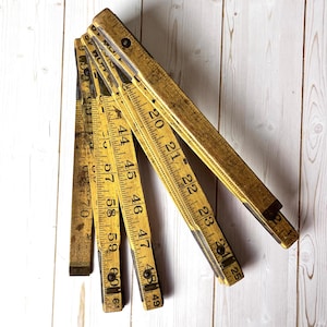 May include: A vintage wooden folding ruler, featuring black measurement markings against a yellowed wood background. The ruler is partially open, displaying inch measurements from 20 to 61 inches. The wood has a distressed appearance.