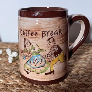 May include: A ceramic coffee mug with a brown rim and handle. The mug features a tan background with a cracked texture and a cartoon illustration of a man and woman dancing. The words "Coffee Break" are written on the mug.