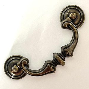 May include: Antique bronze-finished drawer pull with a decorative design. The pull features a curved handle and two circular backplates with screw details. Ideal for cabinets and furniture.