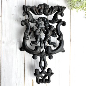 May include: Black cast iron trivet featuring two cherubs, ornate scrollwork, and a decorative bottom. The trivet is designed to protect surfaces from hot cookware and adds a touch of vintage charm to any kitchen.