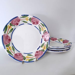 May include: White ceramic plates with a scalloped edge, adorned with a floral design. The plates feature pink roses, green leaves, and yellow accents, outlined with a blue border. A stack of plates is visible.