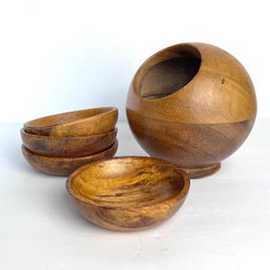 May include: A set of four small, round, brown wooden bowls with visible grain patterns, accompanied by a larger, spherical wooden container featuring a wide, angled opening at the top. The wood displays natural variations in color and texture throughout.