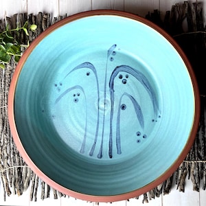 May include: A large, handmade ceramic bowl with a turquoise interior and a reddish-brown rim. The bowl features a hand-painted design of stylized blue plant-like shapes. The bowl is a decorative piece for serving or display.