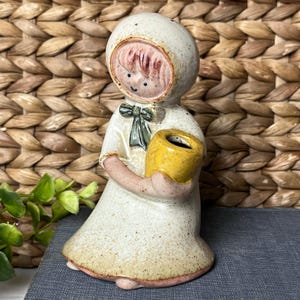May include: A ceramic figurine of a child wearing a hooded cloak and holding a yellow pot. The figurine has a painted face with red hair and a green bow. The cloak is off-white with a speckled texture.