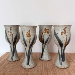 May include: Four handmade ceramic goblets. Each goblet features a speckled, beige finish with painted floral and leaf designs in brown and dark blue. The goblets have a wide base and a flared top, suitable for beverages.