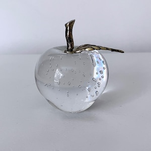 May include: A clear glass apple sculpture with internal bubbles. The apple has a bronze-colored stem and a leaf. The sculpture is a decorative item, likely for display on a shelf or table.