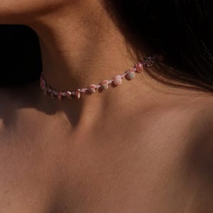 May include: A delicate pink seashell choker necklace. The necklace features small, natural seashells and beads strung together on a thin, light-colored cord. The shells vary in size and shape, creating a unique and organic design.