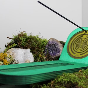 Può includere: Un porta incenso in legno verde con un disegno a spirale sul retro. Il supporto è circondato da muschio, cristalli e piccoli fiori gialli. Un bastoncino di incenso nero è inserito nel supporto.
