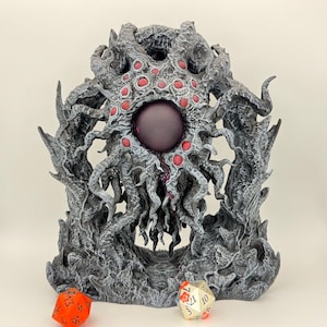 May include: A gray, tentacled sculpture with a central purple orb and red accents. Two multi-sided dice are in the foreground. The sculpture has a detailed, textured appearance, suggesting a fantasy or tabletop gaming theme.