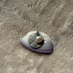 May include: A silver-toned pendant featuring a circular, hammered metal disc layered with a single, off-white, irregular freshwater pearl. The pendant is attached to a small silver jump ring, creating a layered, textured appearance.