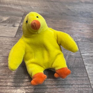 May include: A plush yellow duck toy with orange feet and beak. The duck has small black eyes and outstretched arms. The toy is made of a soft, fuzzy material.