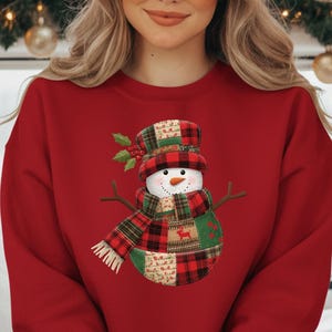 Könnte beinhalten: Ein rotes Sweatshirt mit einem Schneemann-Design. Der Schneemann hat ein Patchwork-Design mit kariertem Hut, Schal und Körper. Der Schneemann hat Zweigarme und eine Karottennase. Das Sweatshirt ist ein festlicher Weihnachtsartikel.