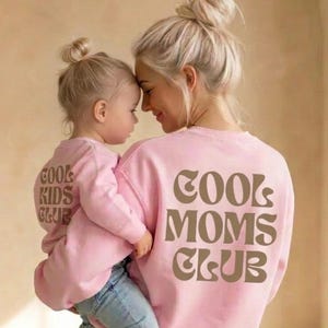 Könnte beinhalten: Eine Mutter und ihr Kind tragen passende rosa Sweatshirts mit dem Text "COOL MOMS CLUB" und "COOL KIDS CLUB" in brauner Schrift. Die Mutter hält das Kind, beide tragen blaue Jeans.