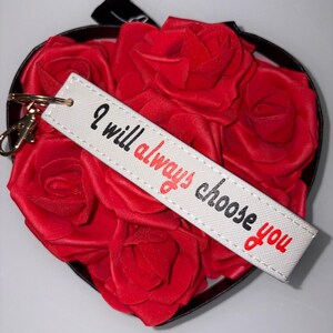 Könnte beinhalten: Eine herzförmige Schachtel gefüllt mit roten Rosen, mit einem weißen Schlüsselanhänger mit der Aufschrift "I will always choose you". Der Schlüsselanhänger hat einen goldfarbenen Verschluss. Die Rosen sind leuchtend rot, und die Schachtel hat eine schwarze Umrandung.