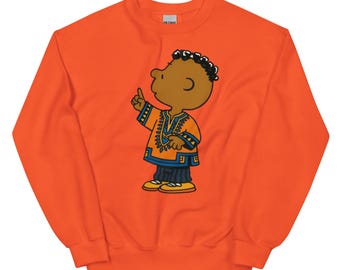 May include: Orange sweatshirt featuring a cartoon illustration of a person in traditional African attire. The character is brown-skinned with black curly hair and is pointing upwards. The outfit is orange and blue. The sweatshirt has a crew neck and long sleeves.