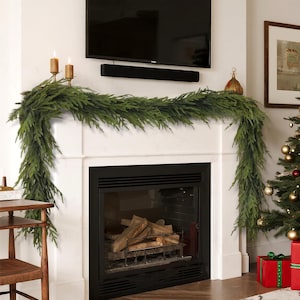 May include: A white fireplace mantel is decorated with a green garland. Two lit candles in gold holders sit on the mantel. A Christmas tree with ornaments and wrapped gifts is to the right of the fireplace.
