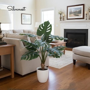 May include: A large, artificial Monstera plant with vibrant green leaves in a white pot. The plant is placed in a living room with hardwood floors, a beige sofa, and a fireplace. The image includes the text "Silvanest".