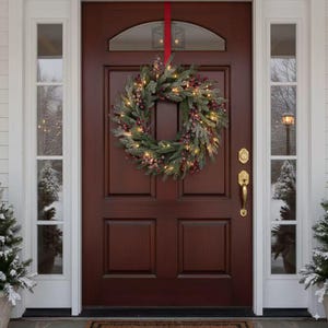 May include: A circular wreath made of frosted green pine needles and red berries, illuminated by small warm white lights. The wreath is hung by a red ribbon on a dark brown wooden front door with glass side panels and gold hardware.