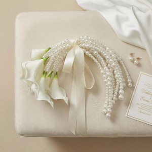 May include: A bridal bouquet featuring white calla lilies and cascading strands of faux pearls. The bouquet is tied with a cream-colored satin ribbon. Gold stud earrings and a wedding invitation are also visible.